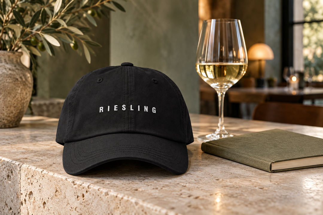 Black Riesling cap with white embroidery on a pale stone wine bar beside a wine glass and closed notebook.
